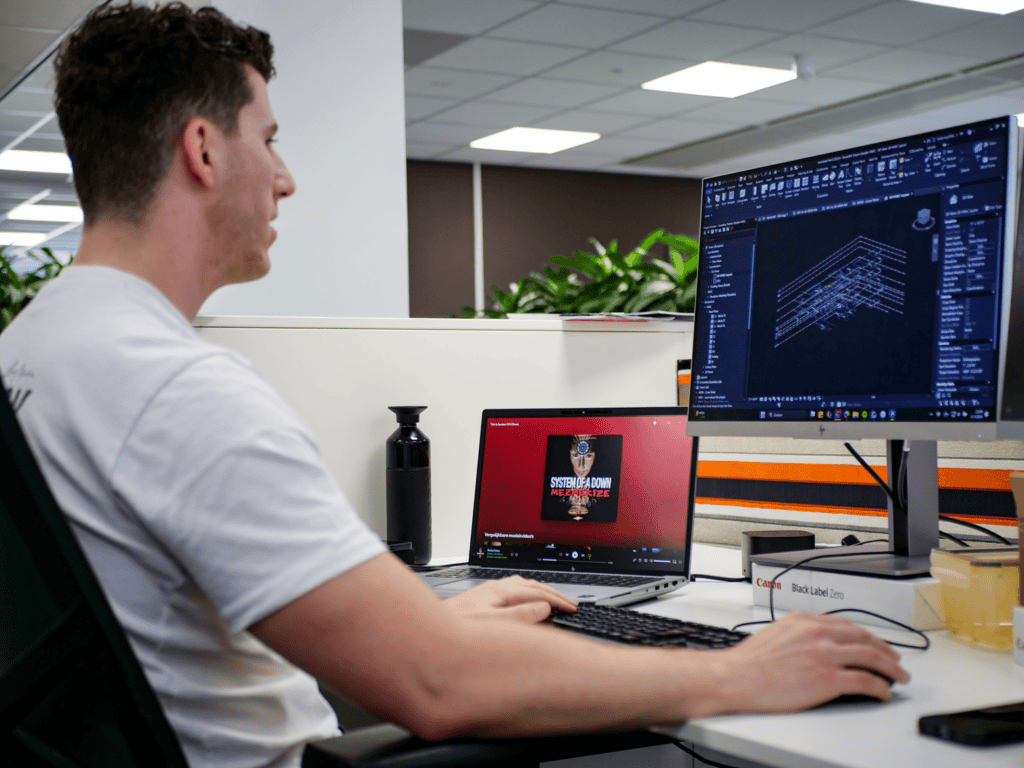 Een jonge engineer werkt geconcentreerd aan een BIM-ontwerp op een moderne werkplek in een kantooromgeving. Op een extern beeldscherm is technische ontwerpsoftware zichtbaar met een gedetailleerd 3D-model, terwijl op de laptop muziek wordt afgespeeld. De werkplek is strak ingericht met een toetsenbord, muis en accessoires, wat een professionele en productieve werksfeer benadrukt. Op de achtergrond zorgen planten en een open kantoorindeling voor een frisse, hedendaagse uitstraling.