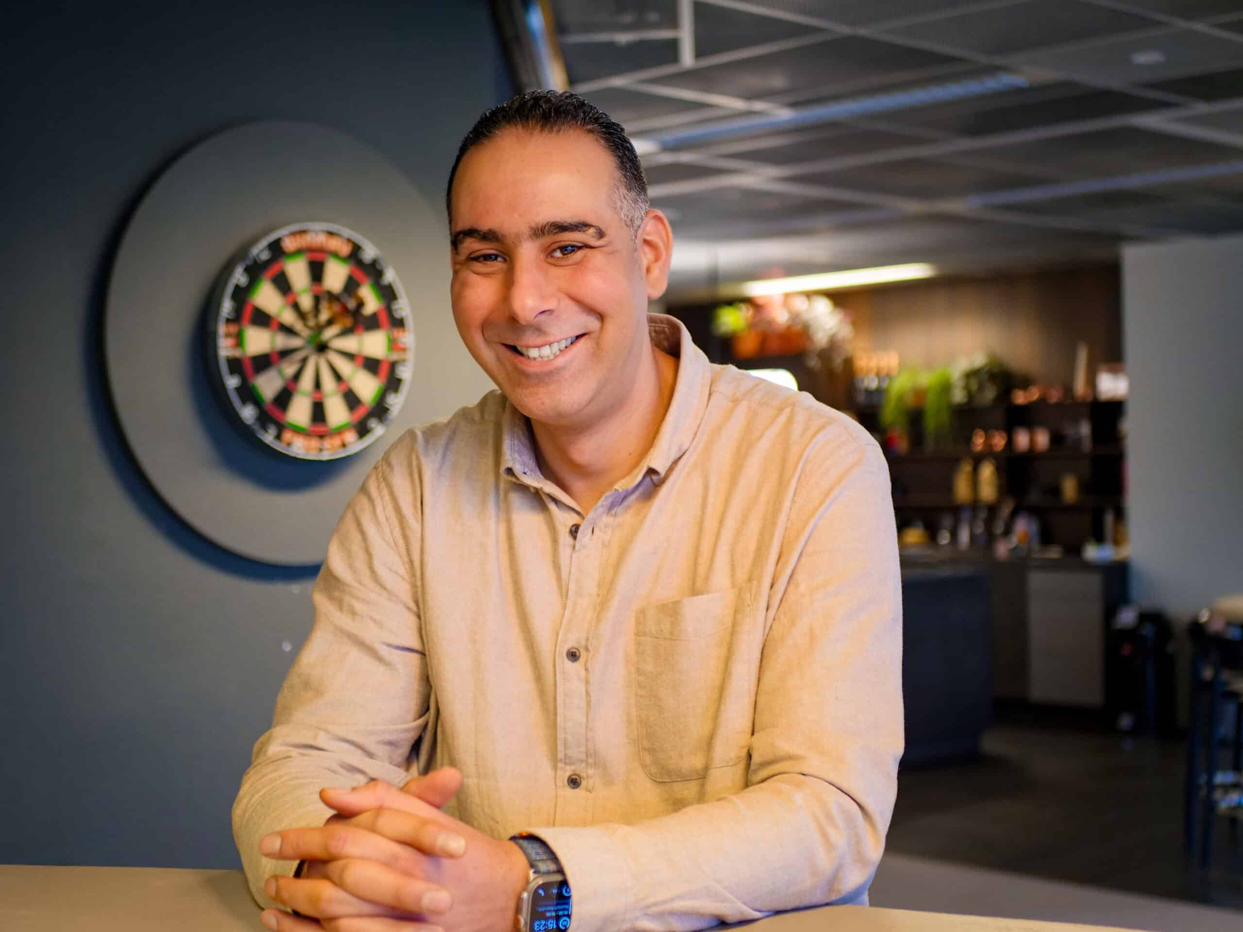 Youssef is beveiligingsexpert bij Bluetech Engineering. Hij staat met z'n handen in elkaar gesloten en een fijne glimlach. Op de achtergrond zie je een dartbord en de keuken van het hoofdkantoor in Leusden.