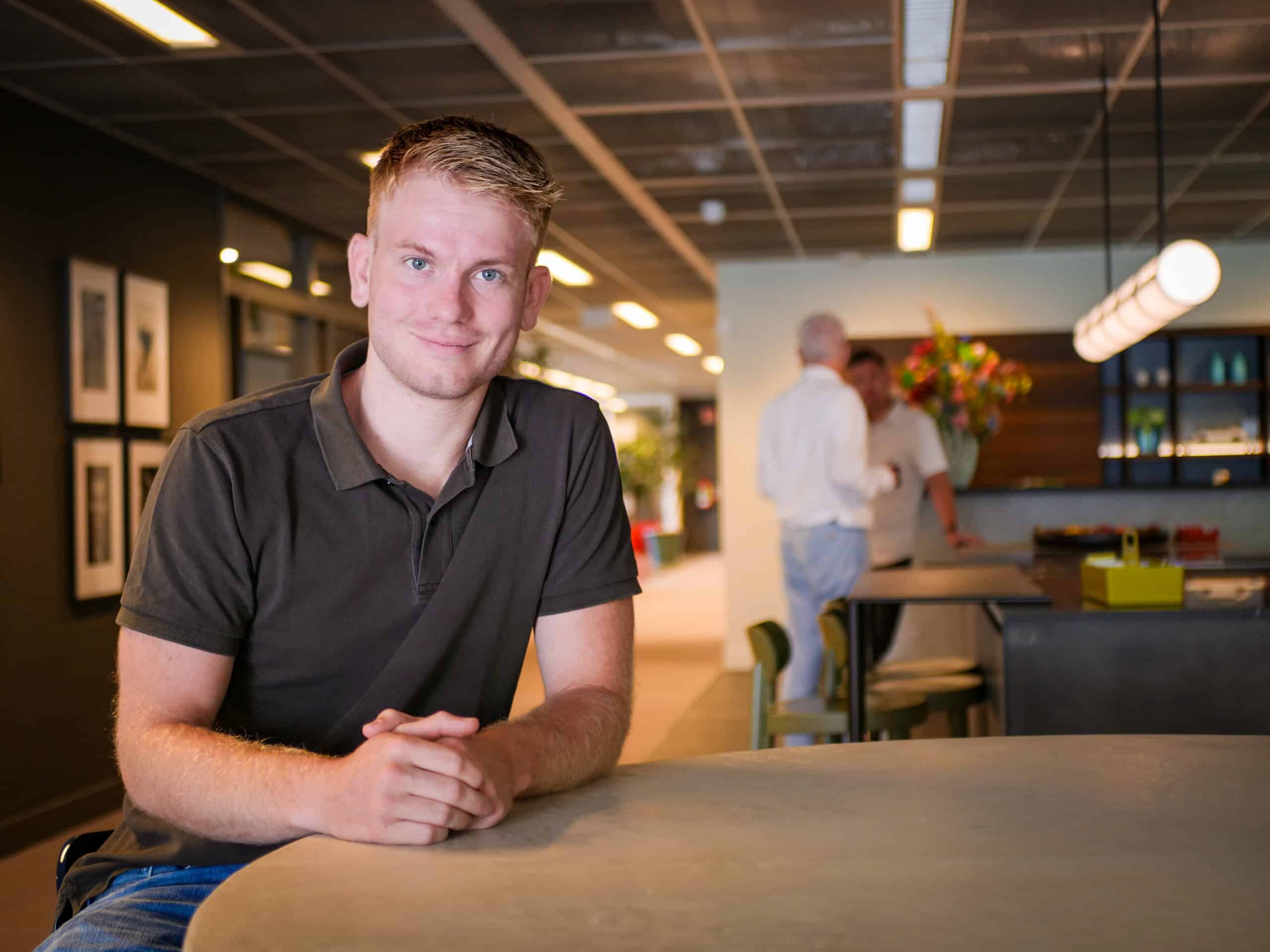 Marc Kooistra is business consulent en is gedetacheerd vanuit Bluetech Engineering. Je ziet hem staan op ons hoofdkantoor in Leusden. Op de achtergrond zijn mensen in gesprek aan het kookeiland. Marc draagt een donkergrijze polo, heeft blond haar en blauwe ogen.