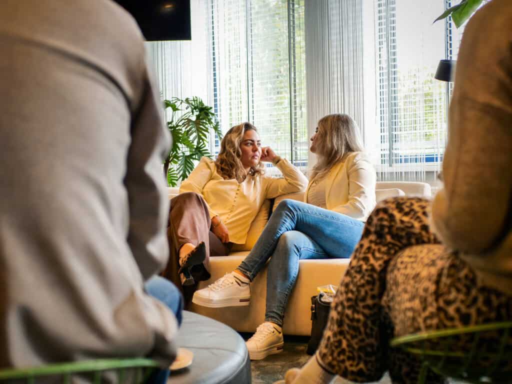 Op de foto zie je 2 personen in gesprek in een informele setting. Ze zitten op de bank waarbij 1 van de twee aandachtig luisterd.